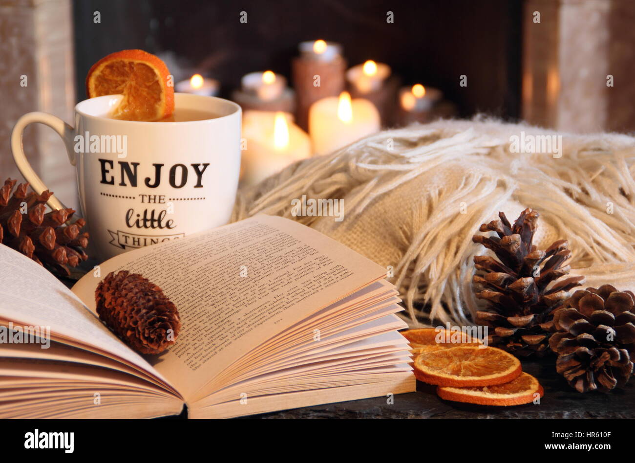 Reading a paperback book by an open fire in a cosy hygge inspired English living room with a hot drink and blanket (throw) on an autumn night Stock Photo