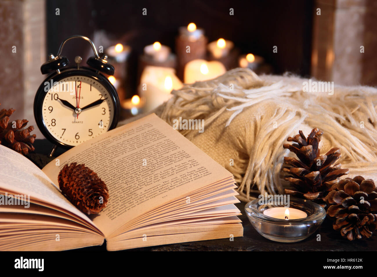 Reading a paperback book at night by candle light next to an open fireplace in a cosy English home with a blanket (throw) in winter Stock Photo