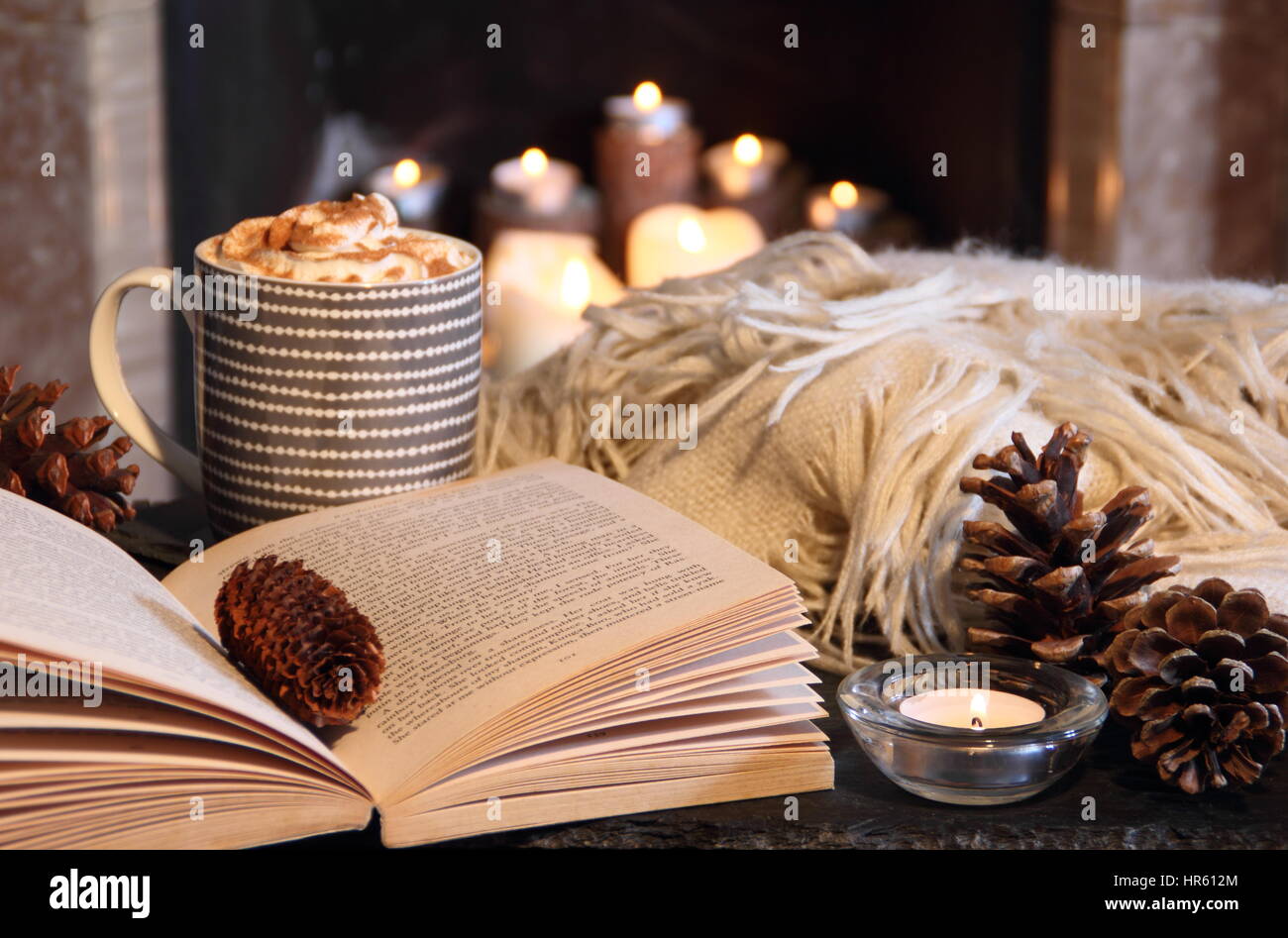 Reading a paperback book by candle light next to an open fireplace in a cosy English home with a creamy hot drink and blanket on a winter evening Stock Photo