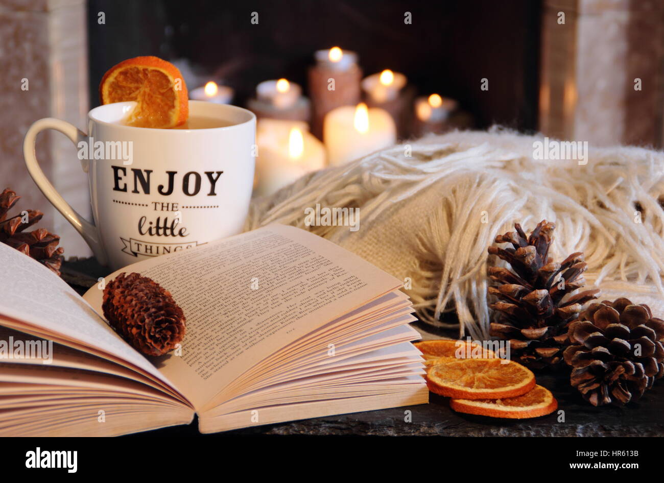 Reading a book by an open fireplace in a cosy hygge inspired English living room with a hot drink and blanket (throw) as autumn turns to winter Stock Photo