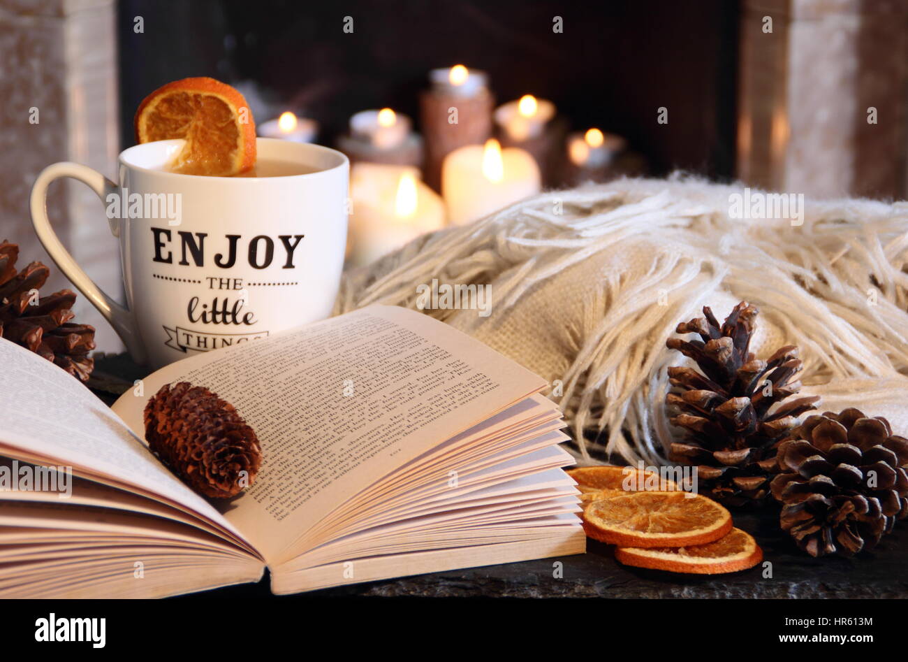 Reading a paperback book by an open fireplace in a cosy hygge inspired English living room with a hot drink and blanket (throw) - autumn/winter Stock Photo