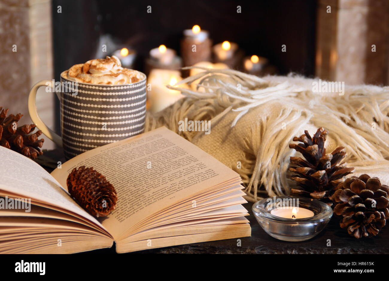 Reading by a fireplace by candle light in a cosy English home with a creamy hot drink and blanket (throw) in late autumn/early winter Stock Photo