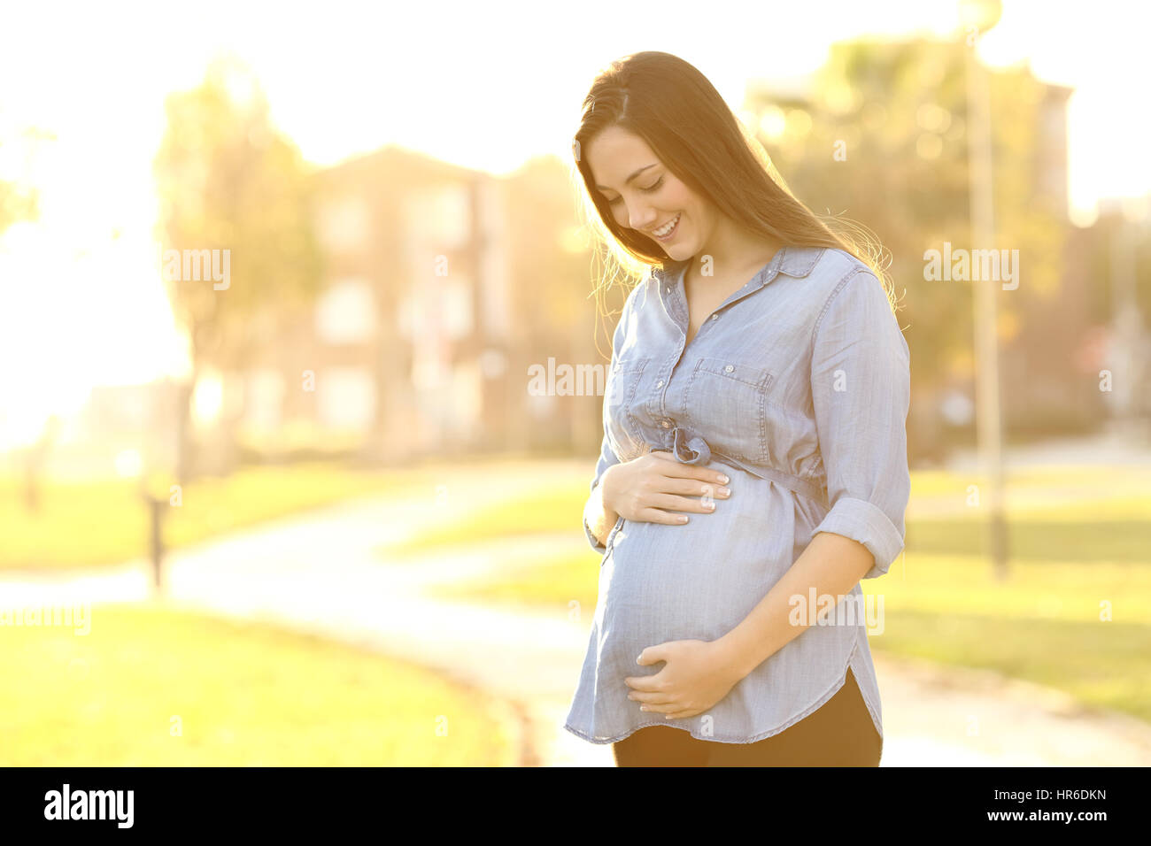 Portrait of a happy and proud pregnant woman looking at her belly in a park at sunrise with a warm back light in the background Stock Photo