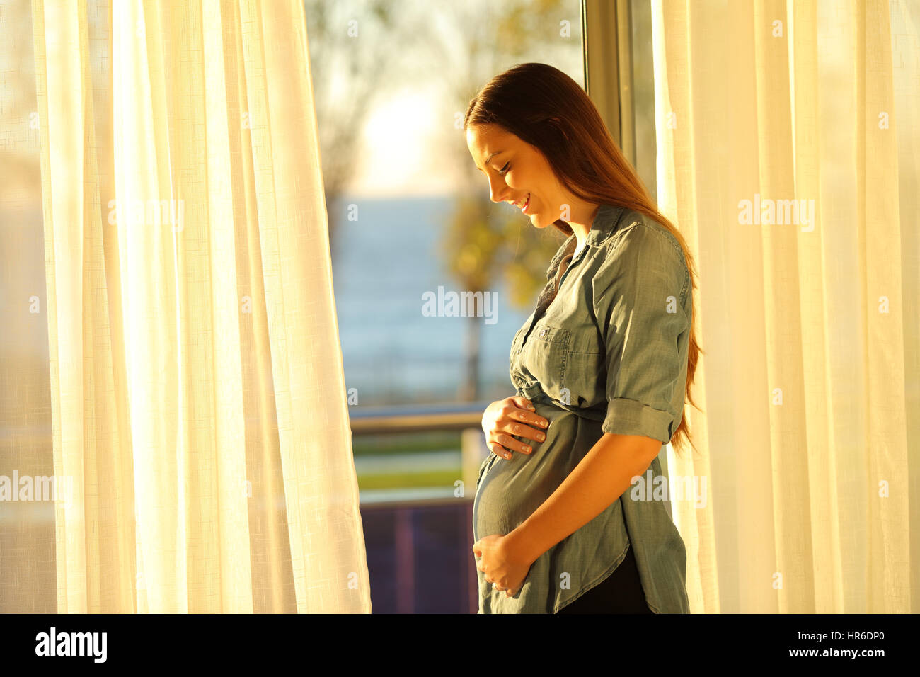 Pregnant woman watching her belly near a window with a warm sunset light at home Stock Photo