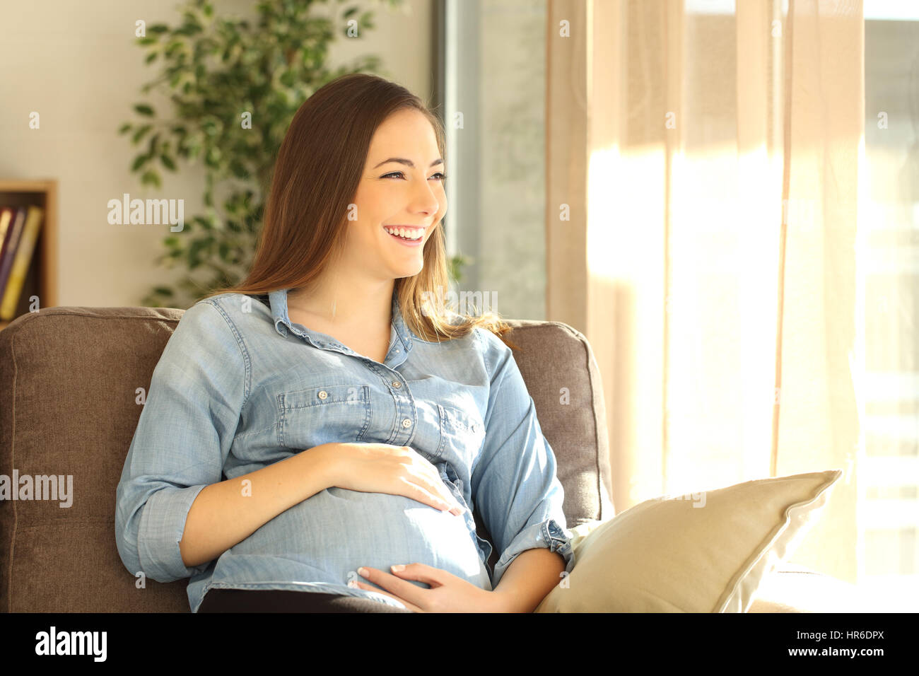 Pregnant woman looking through a window sitting on a sofa in the living room at home with a warm sunlight Stock Photo