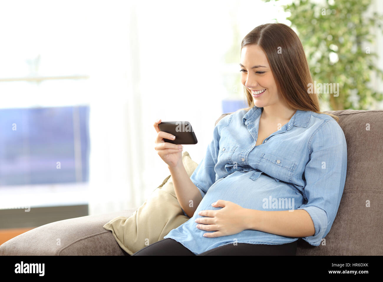 Pregnant woman watching media content in a phone sitting on a couch in the living room at home Stock Photo