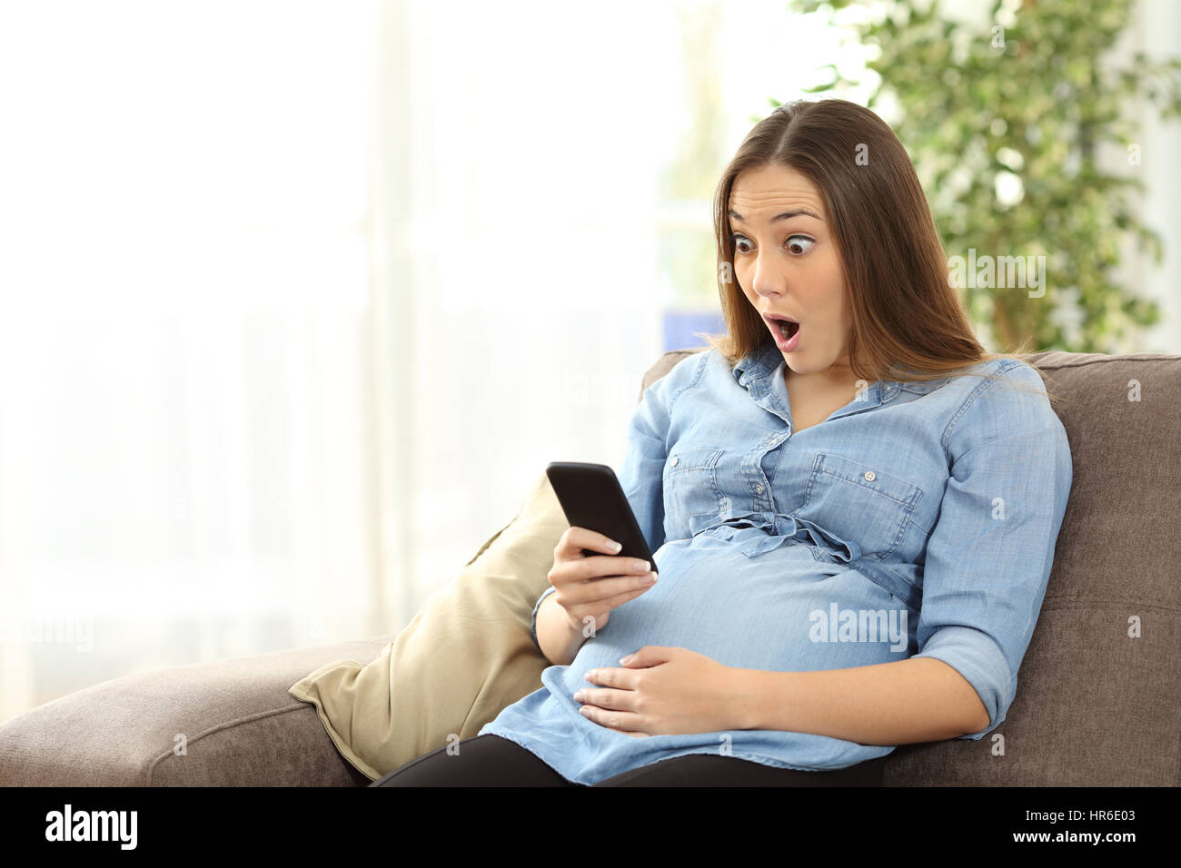 Amazed pregnant woman watching media content in a smart phone sitting on a sofa in the living room in a house interior Stock Photo