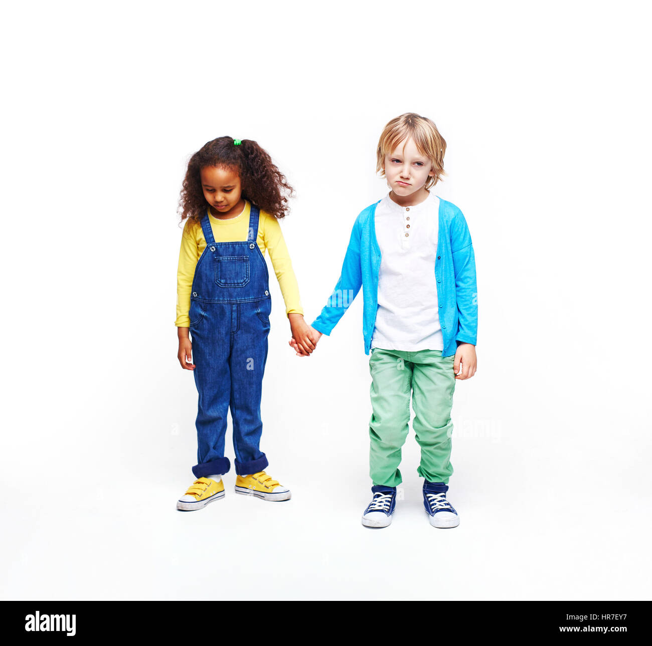 Studio portrait of children against white background: full body shot of two kids, African girl and blond boy, standing together holding hands both loo Stock Photo