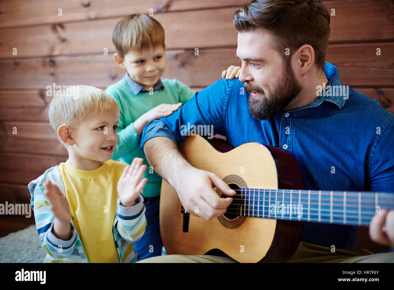 Bearded nursery teacher giving music lesson to little boys: he playing guitar and singing funny song while his pupils listening to him with interest Stock Photo