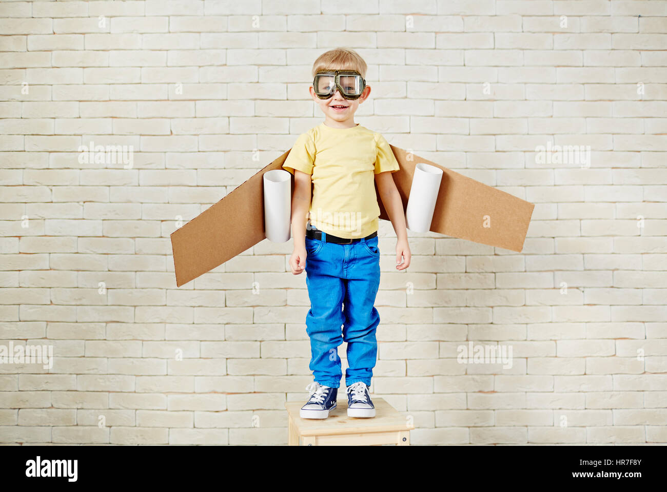 Funny little boy in aviator goggles and with carton airplane wings behind his back standing on wooden chair and looking at camera with smile Stock Photo