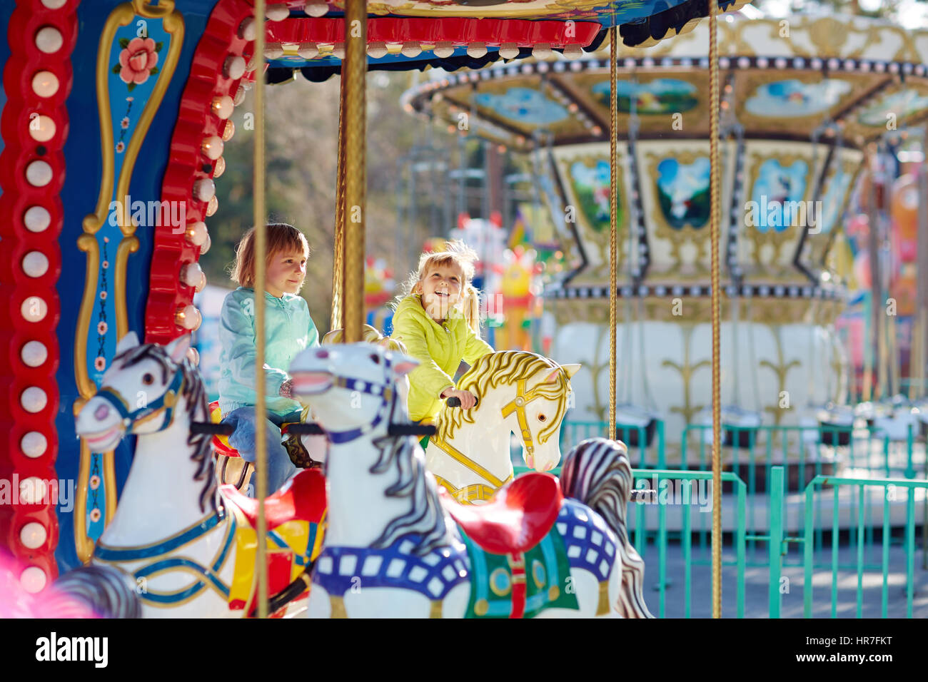 Two blondhaired little girls in bright windbreakers sitting on carousel horses and smiling