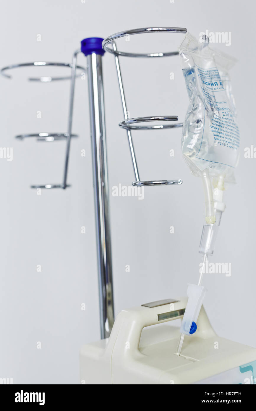 Closeup still life shot of drip bottle with transparent sodium chloride solution hanging on metal stand in hospital ward against white walls Stock Photo