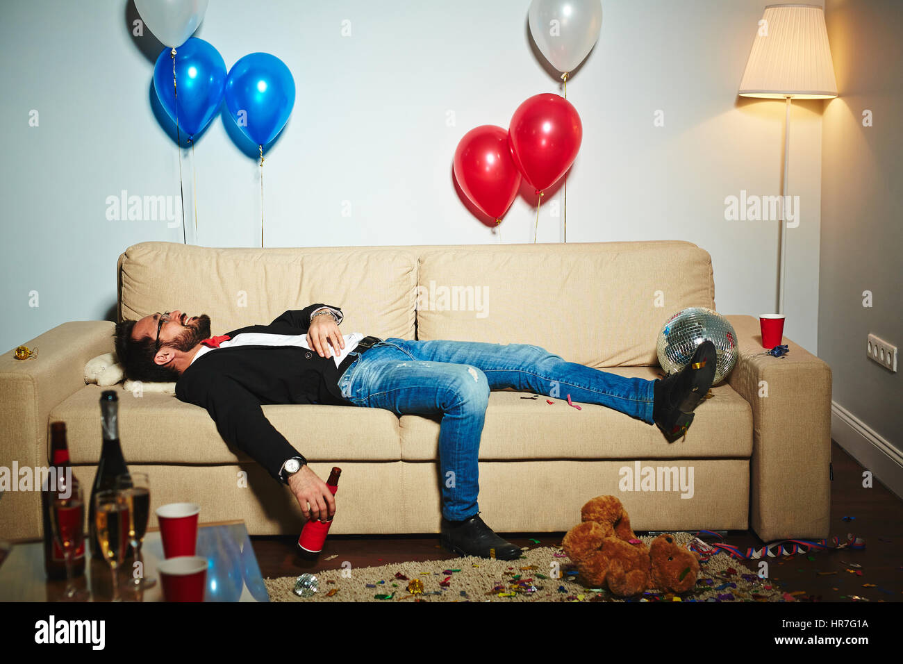 Smiling middle-aged man lying on couch and drinking beer from bottle in messy living room, empty alcohol bottles and plastic cups standing everywhere Stock Photo