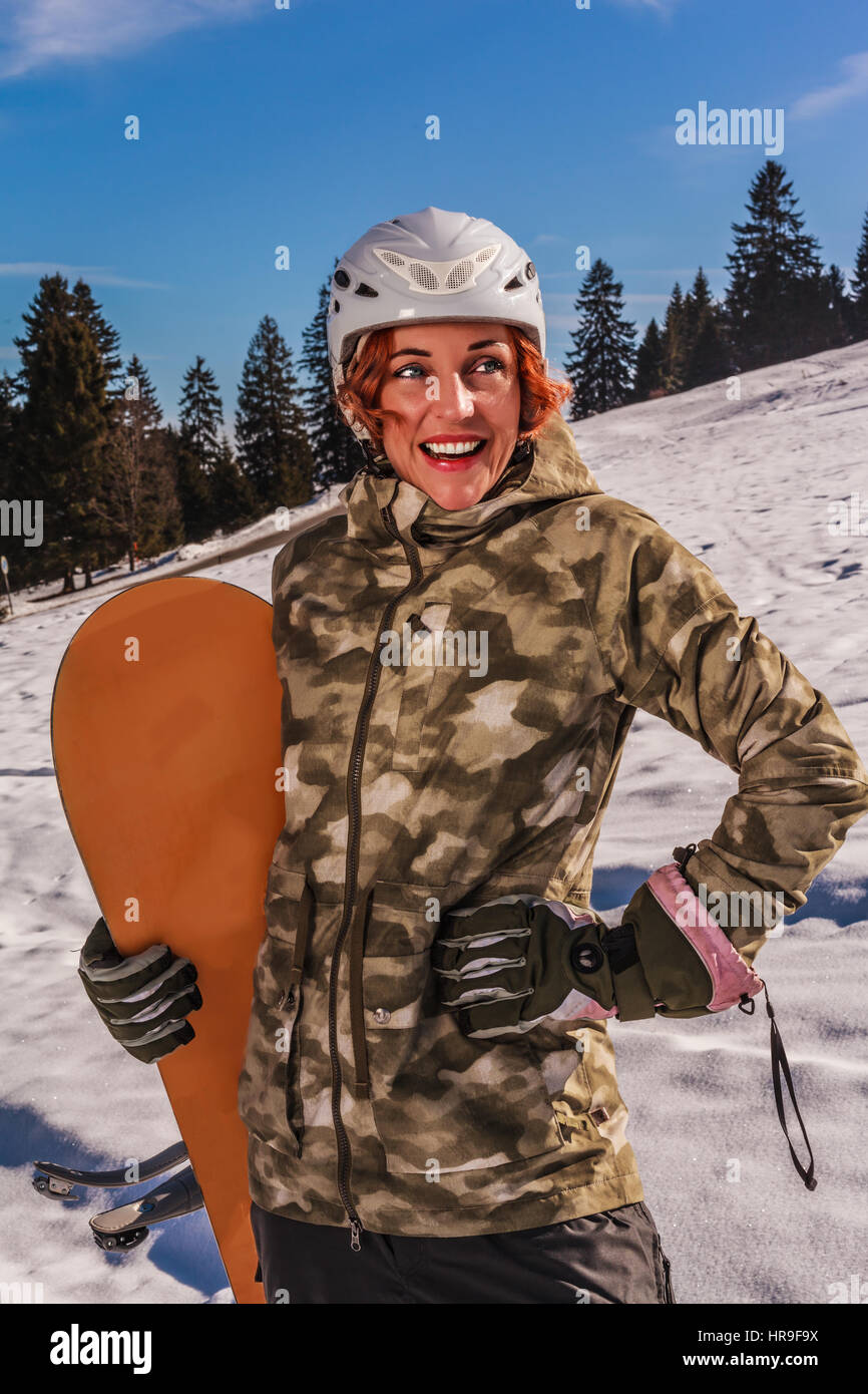 Upper body intake in sunny weather of a redheaded, laughing woman with helmet, standing on the piste with snowboard in his hand. Stock Photo