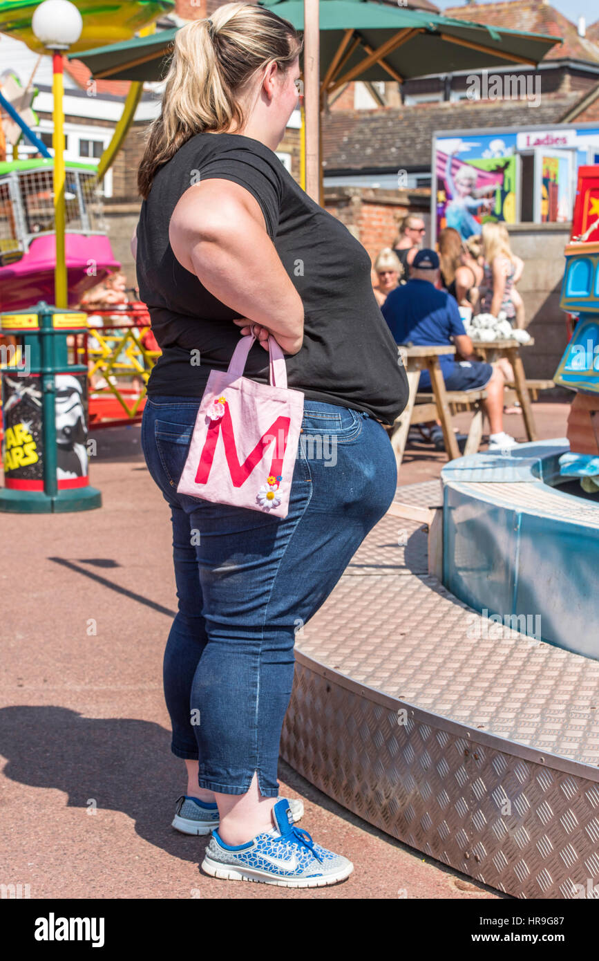 An overweight woman  standing beside a ride at a fairground holding a small child's pink bag. Stock Photo
