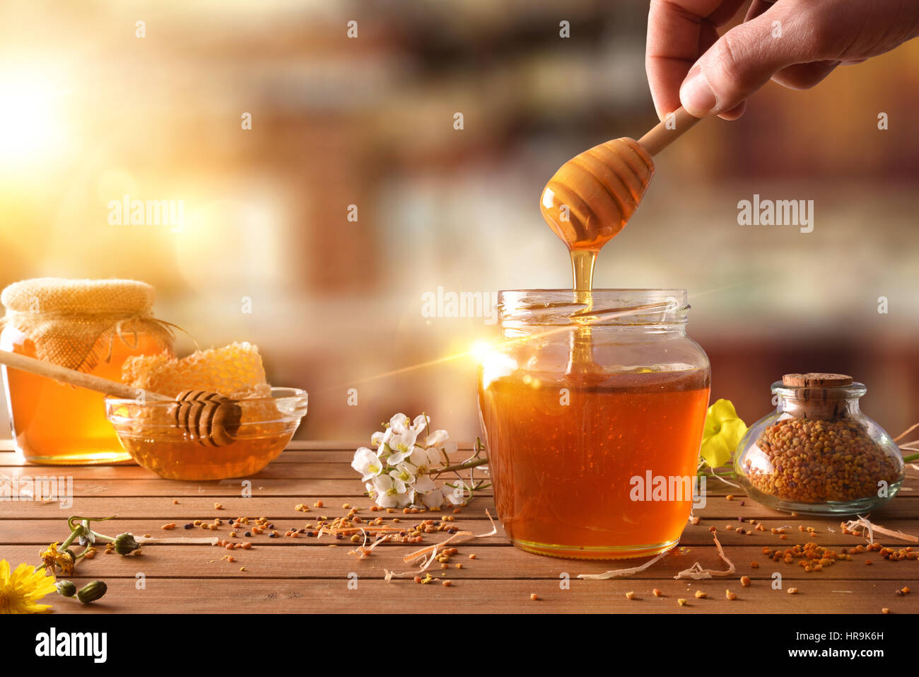 Hand with dipper picking honey from a jar of honey. Jars of honey, bee honeycomb and bee pollen on wooden table with flowers in a rustic kitchen and s Stock Photo