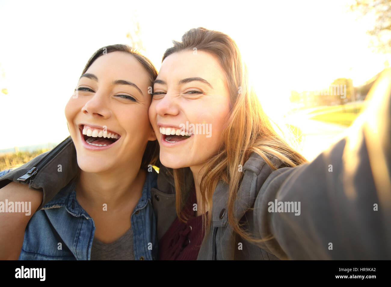 Two funny friends taking selfies outdoors in the street at sunset with a warm light in the background Stock Photo