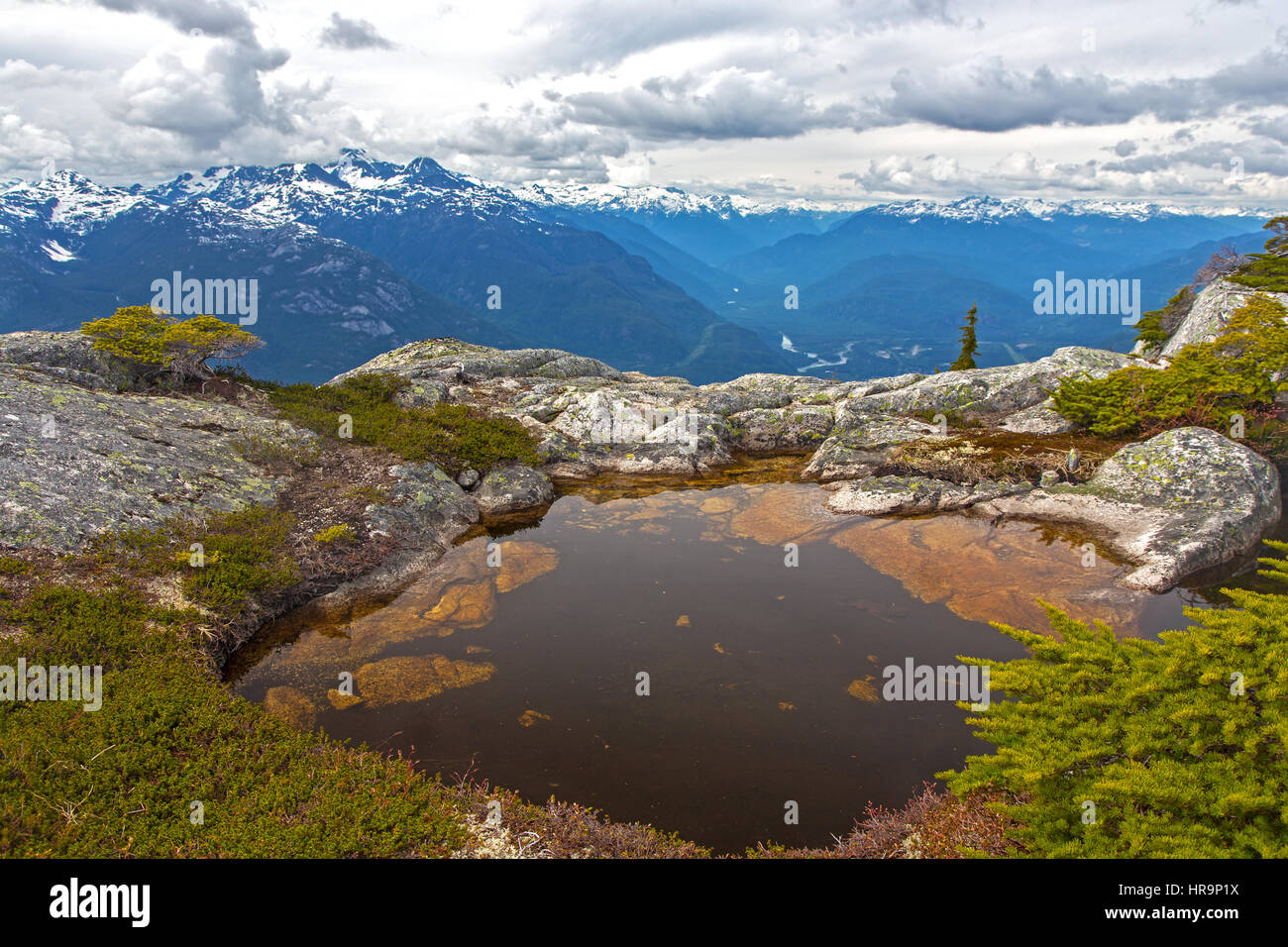 Small alpine lake in rock fissure with distant snowy Tantalus mountains and dramatic cloudy sky near Squamish Coast Mountains British Columbia Canada Stock Photo