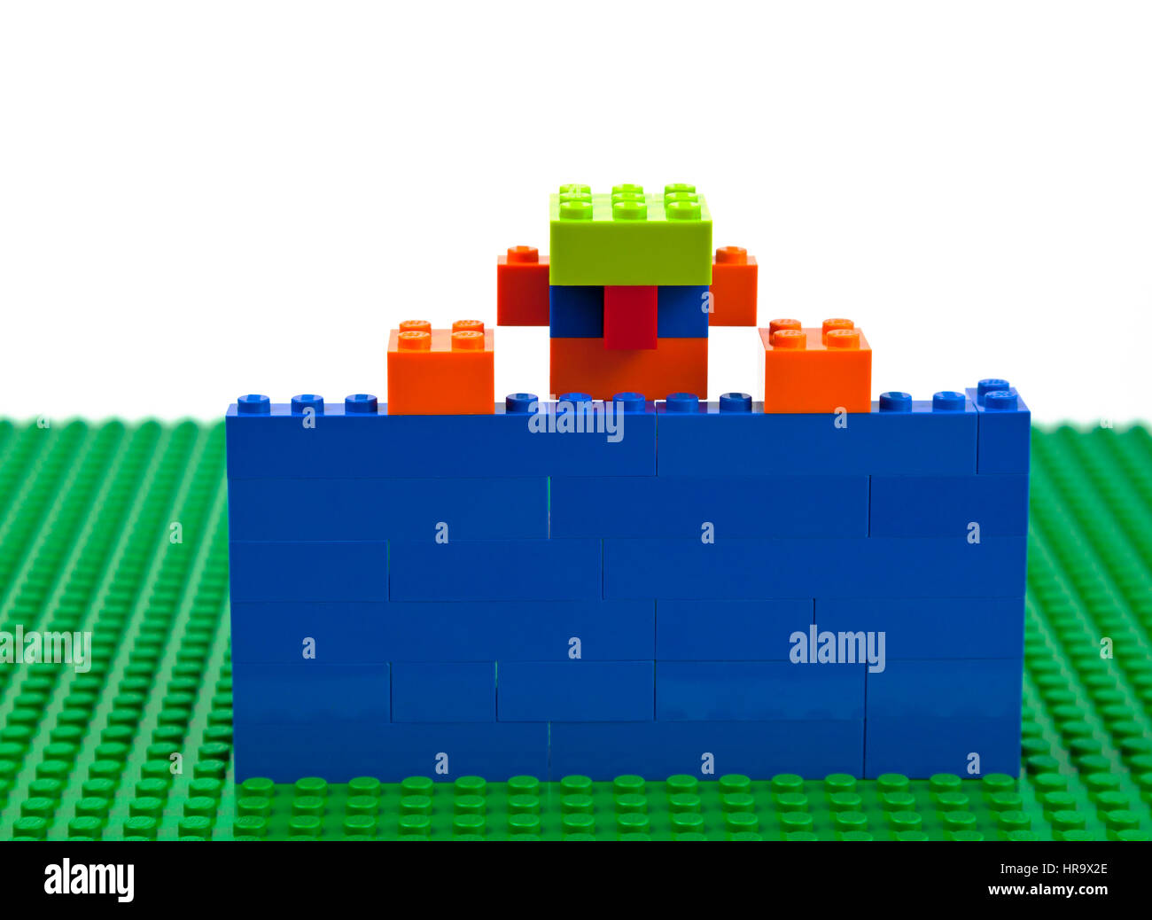 Lego brick construction of a boy, man, male with jug-ears and a green cap confined behind a blue wall raising himself to flee or just saying hello. Stock Photo