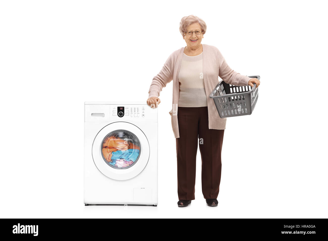 Full length portrait of an elderly woman with an empty laundry basket standing next to a washing machine isolated on white background Stock Photo