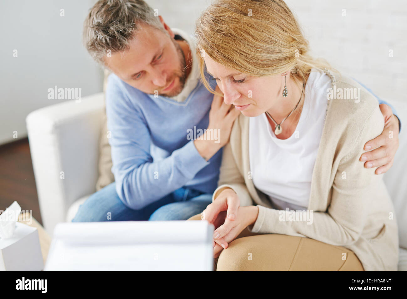 Upset blond-haired woman with tears on her cheeks sitting next to her psychologist while he trying to comfort her Stock Photo