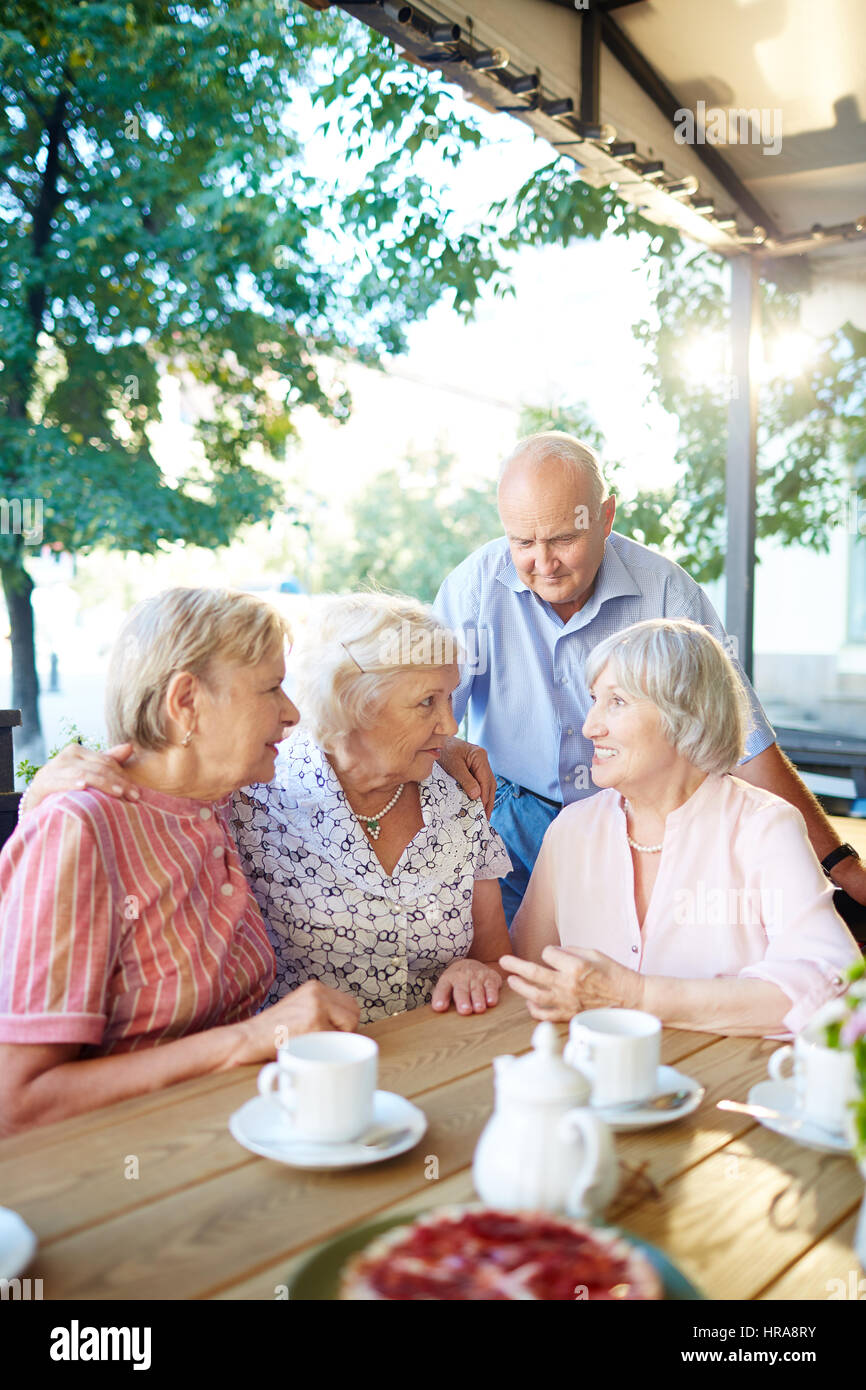 Senior friends having tea party in lovely summer cafe and sharing latest news with each other, two women putting arms on each others shoulders Stock Photo