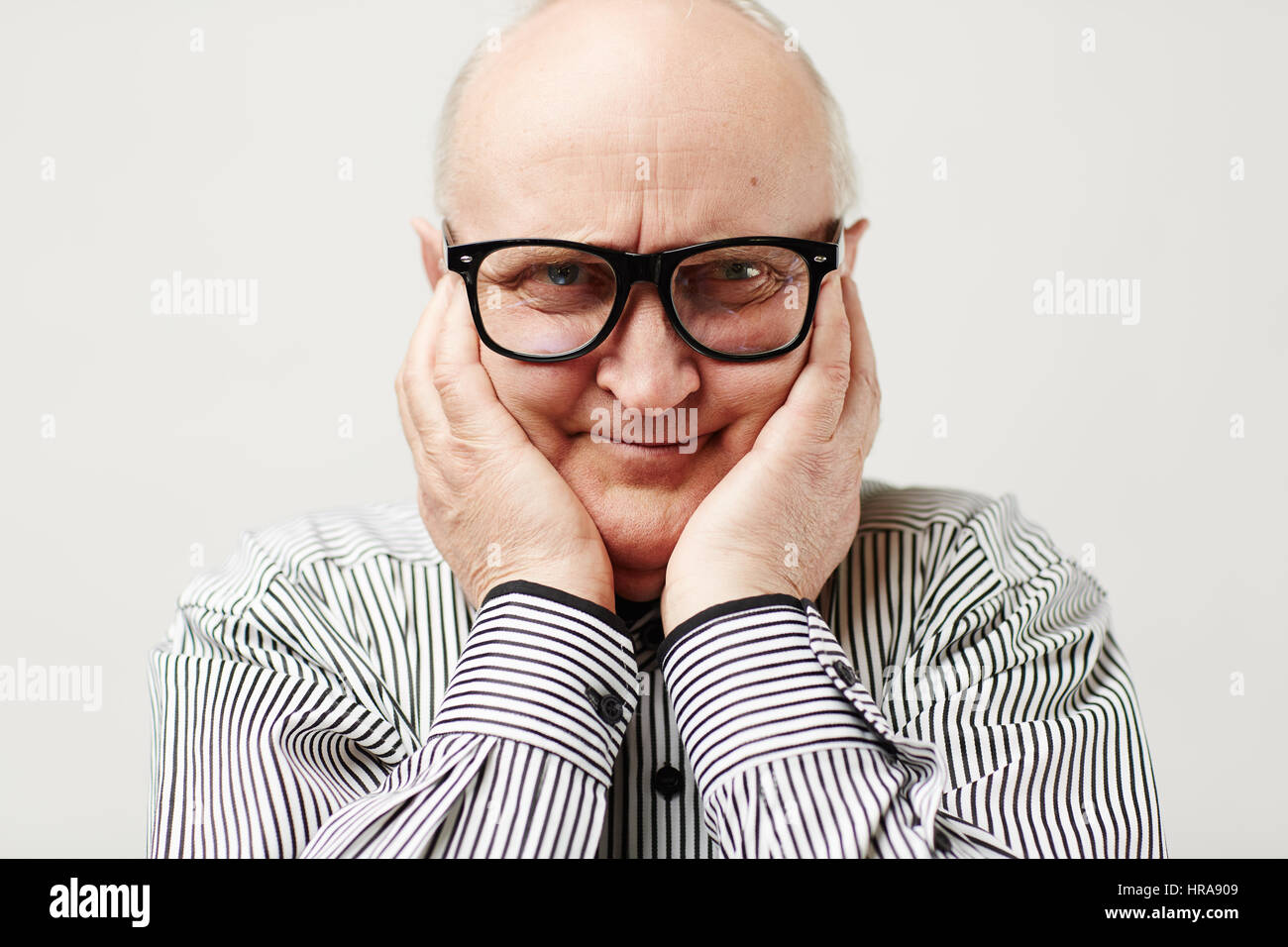 Portrait of charming aged man in eyeglasses and striped shirt making funny face by squishing cheeks with his hands Stock Photo