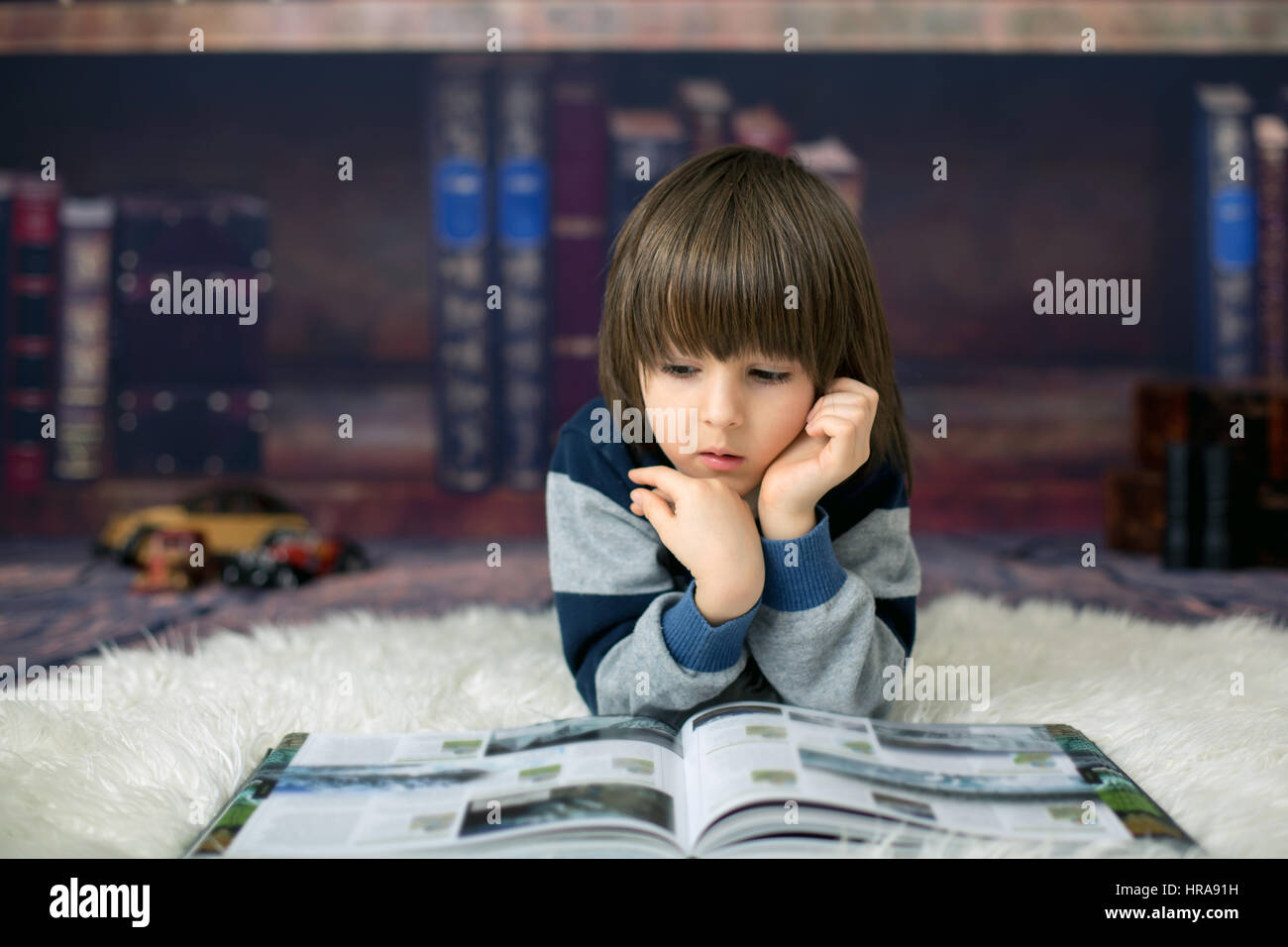 Cute child, boy, reading a book at home in front of the library, lying down on the floor Stock Photo