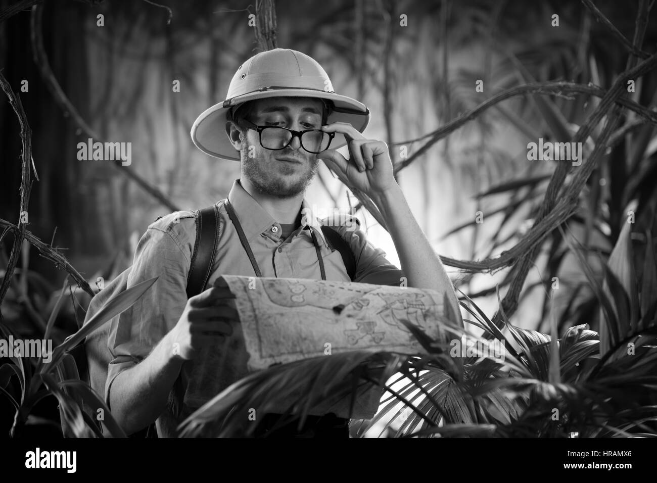 Young confident explorer in the jungle examining an old map and adjusting glasses. Stock Photo