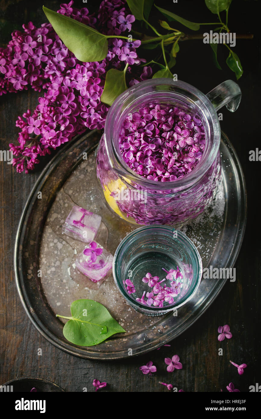 Glass and pitcher of lilac lemonade water with lemon, ice cubes with lilac flowers and lilac branch on vintage iron tray over black wooden table. Dark Stock Photo