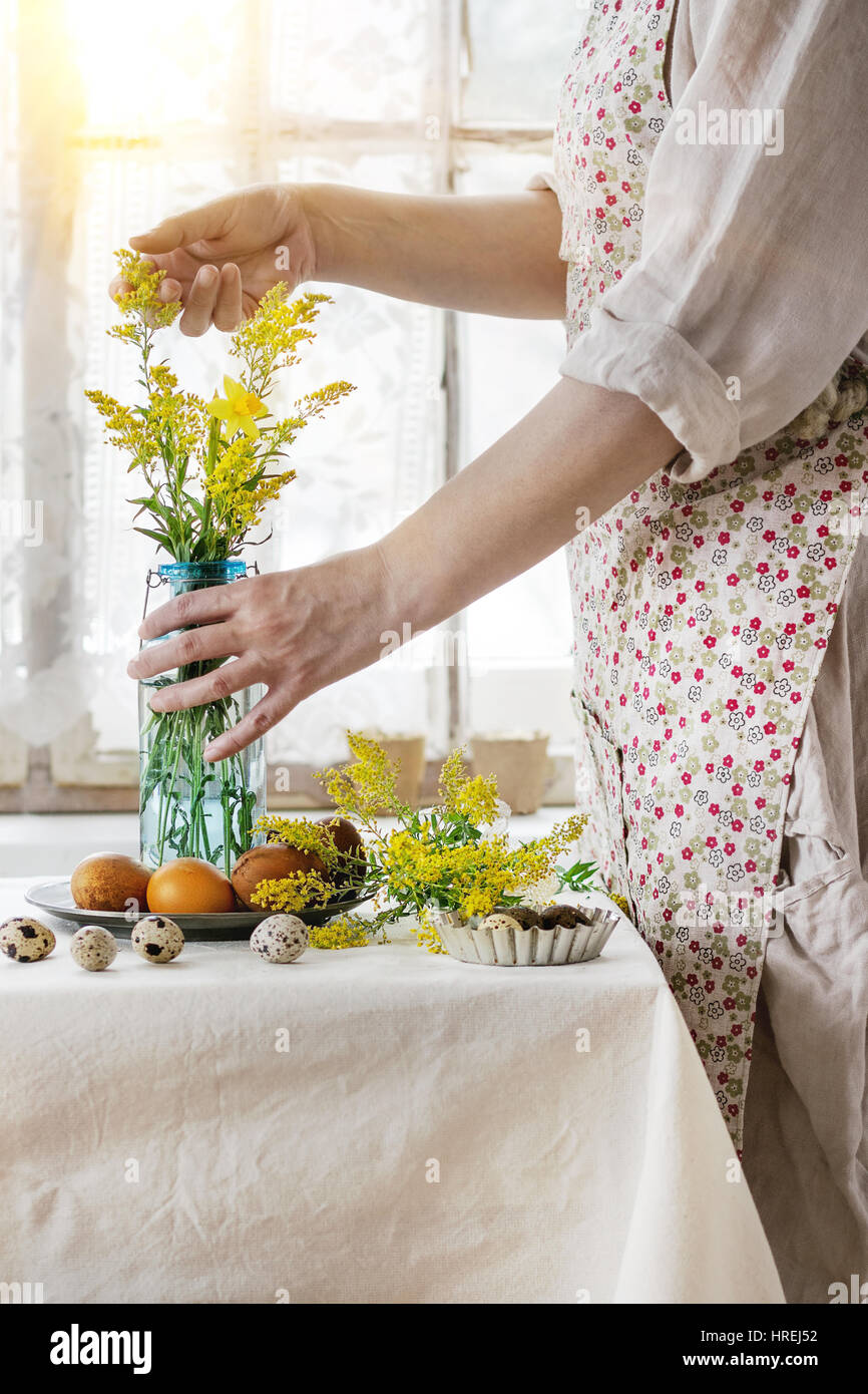 Preparing for Easter. Female hands holding yellow flowers near white tablecloth table decorated brown chicken and quail colored easter eggswith window Stock Photo