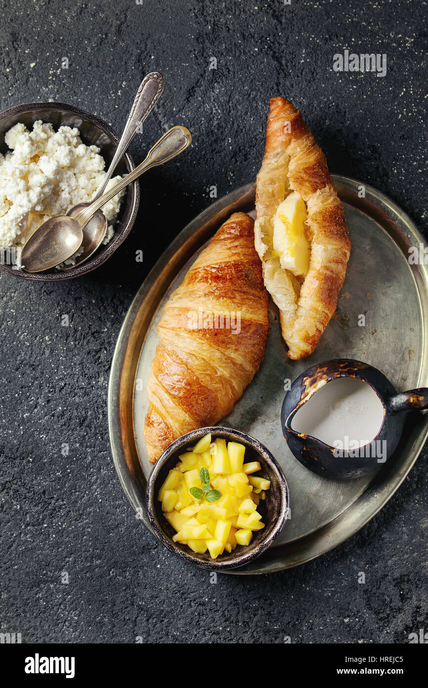 Breakfast with two croissant, butter, cottage cheese, cream and sliced mango fruit, served on serving on metal tray over black concrete texture backgr Stock Photo