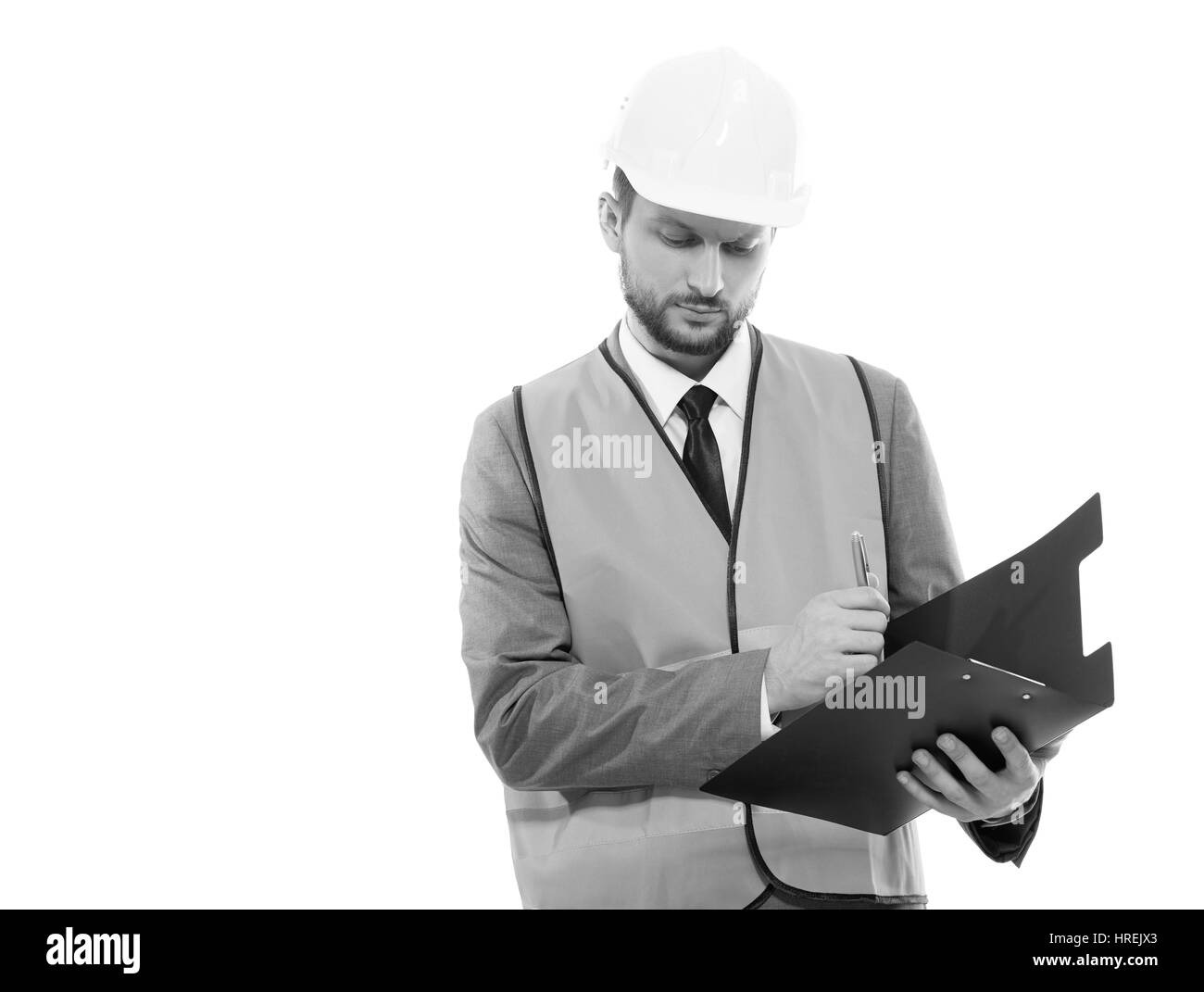Successful developer. Monochrome studio shot of a serious businessman architect wearing protective helmet and safety west writing on his clipboard cop Stock Photo