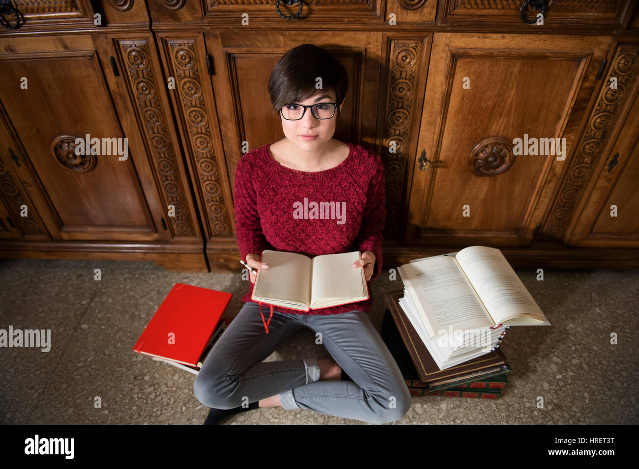 Young female student seated on the floor with open notepad and pencil and with many books around, looking at camera view from the top Stock Photo