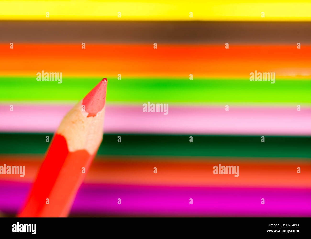 Tip of a red wooden pencil with colored crayons in the background