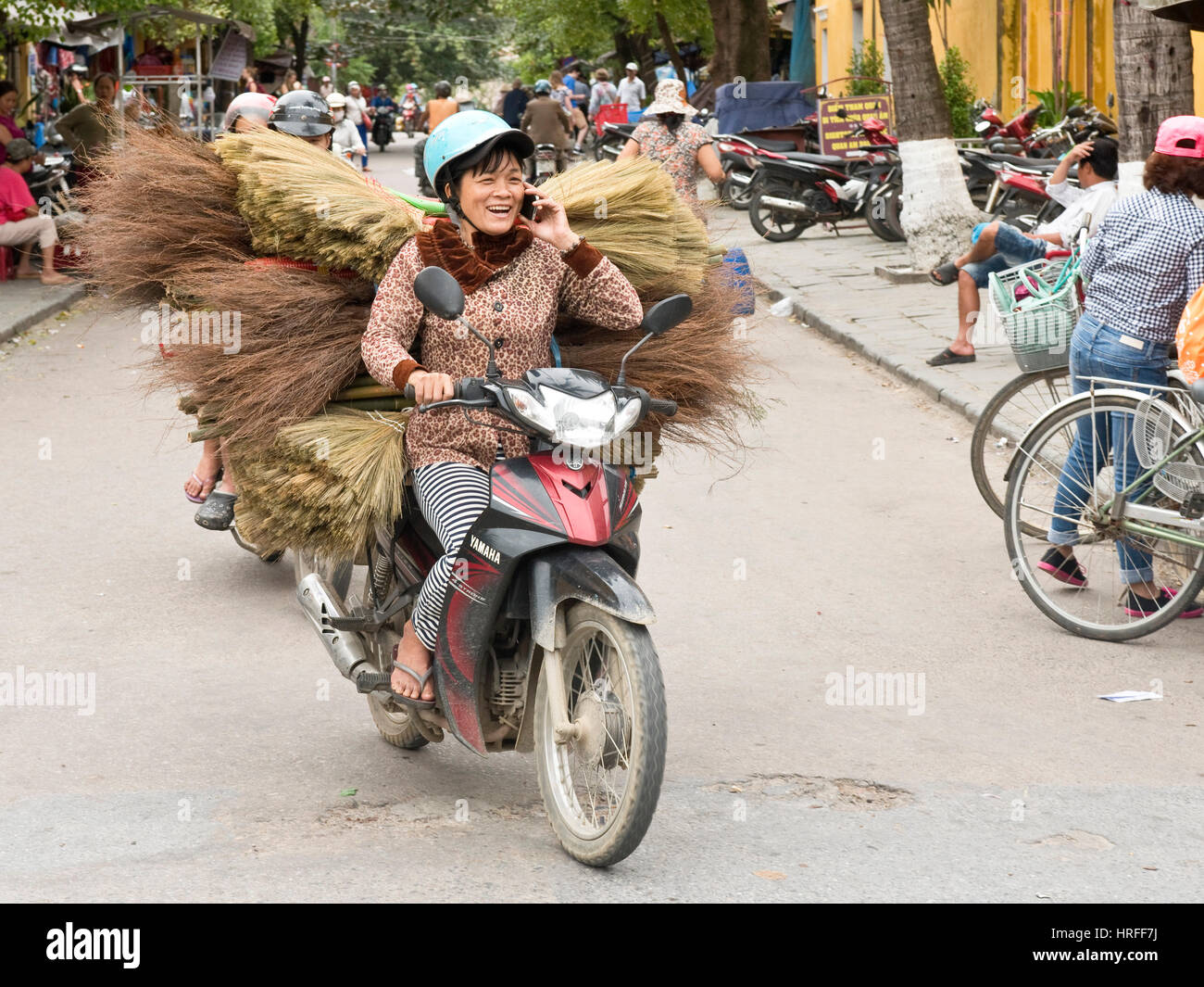 A local vietnamese woman in Hoi An old town riding a moped with a load of brooms while laughing and smiling driving and talking on her mobile phone. Stock Photo