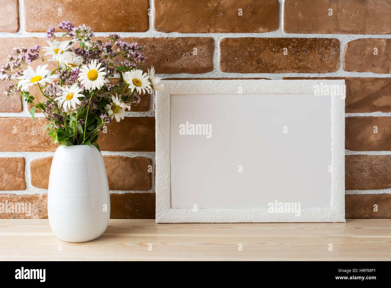 White landscape frame mockup with wwildflowers bouquet in styled vase near exposed brick wall. Empty frame mock up for presentation design.  Template  Stock Photo