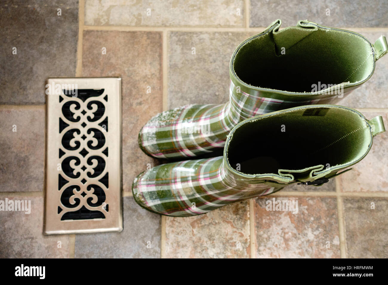 Patterned green women's rubber boots, or Wellies, with snow on the toes warming up at a floor vent indoors. Stock Photo