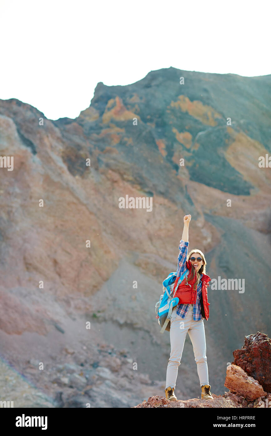 Modern girl in fashionable tourist gear standing on top of peak in mountains raising hand up, inspired and enjoying her freedom Stock Photo