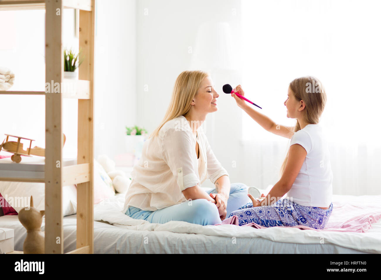 Teenage daughter having fun playing beauty salon with her young blond mother and putting makeup on her face with big puffy brush Stock Photo