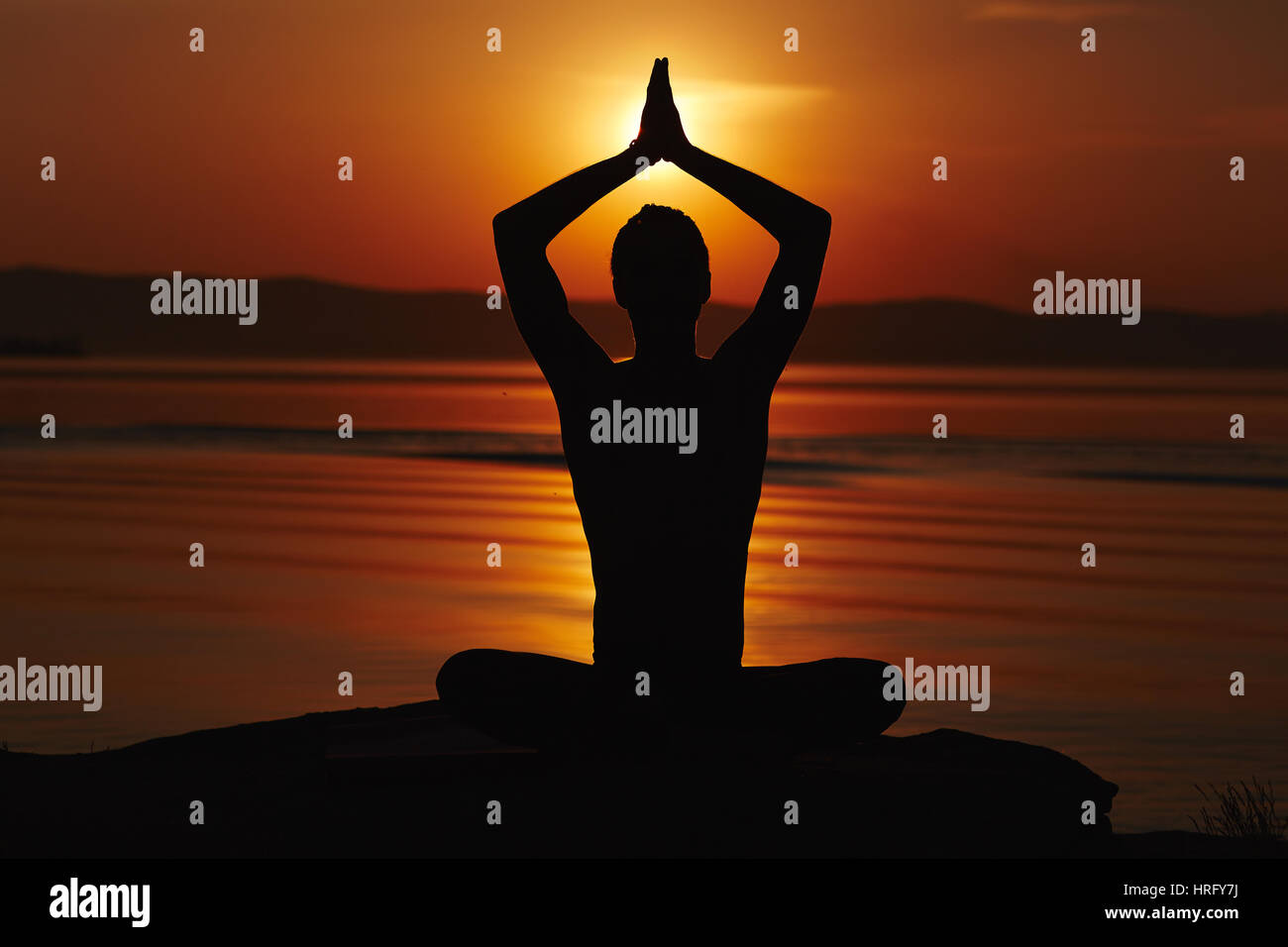 Silhouette of man sitting in lotus pose, holding his arms above head in namaste and meeting sunset Stock Photo