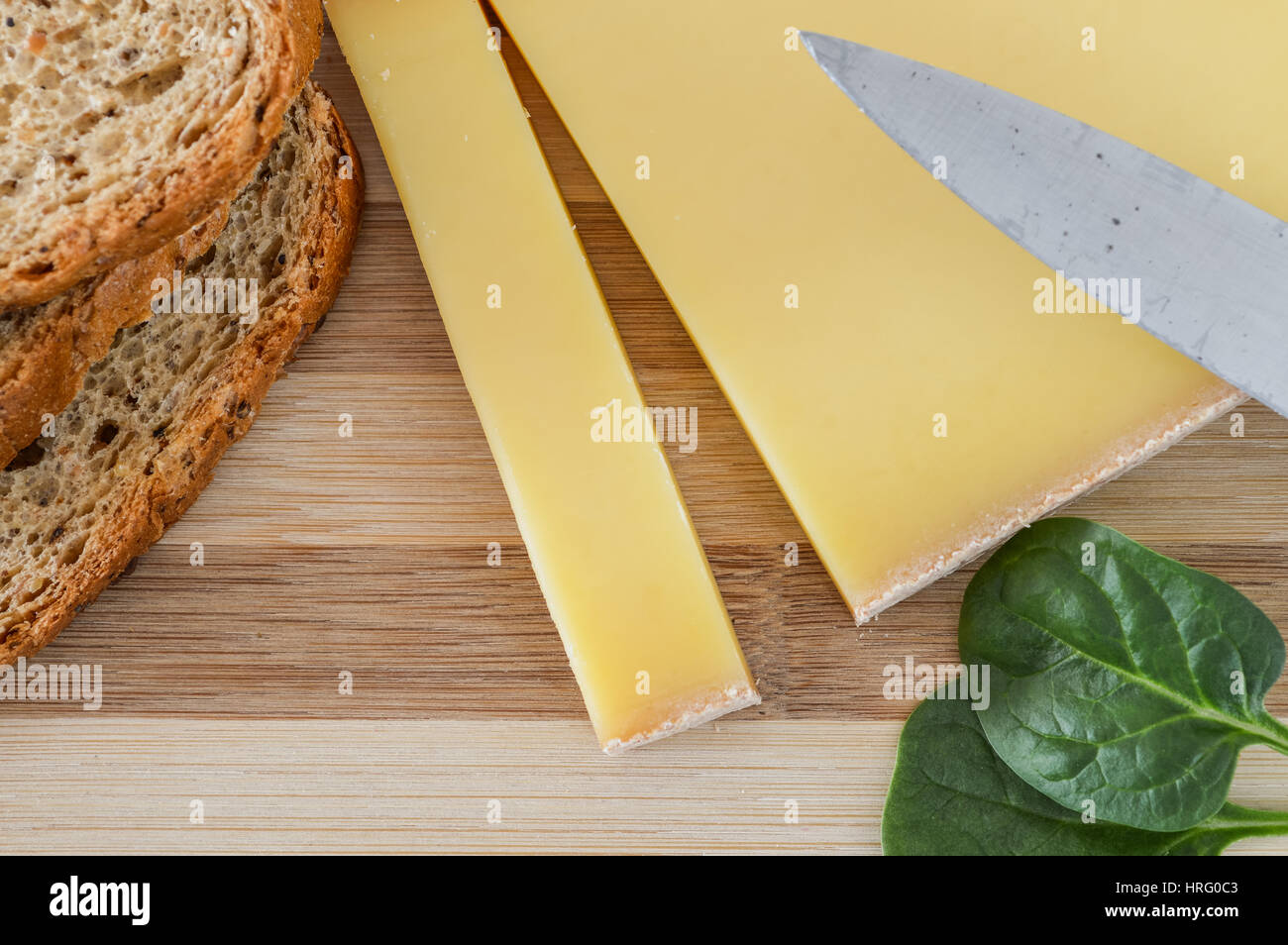 slice of french comte cheese on wood cutting board with fresh green spinach leaves and sliced 7-grain bread closeup Stock Photo