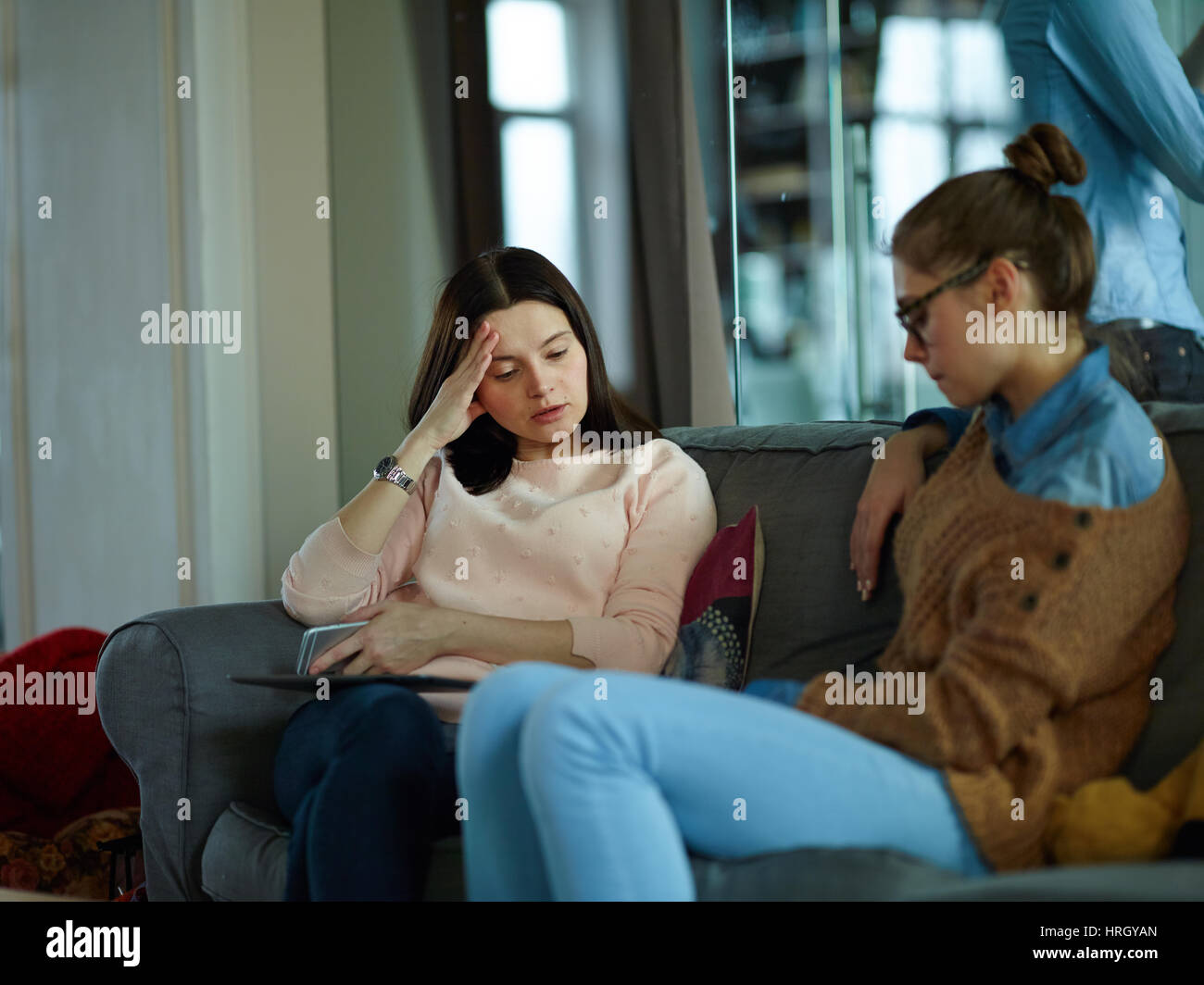 Portrait of two young women on sofa in resting area of dim office, sitting back and looking down tiredly, focus on exhausted brunette woman rubbing he Stock Photo