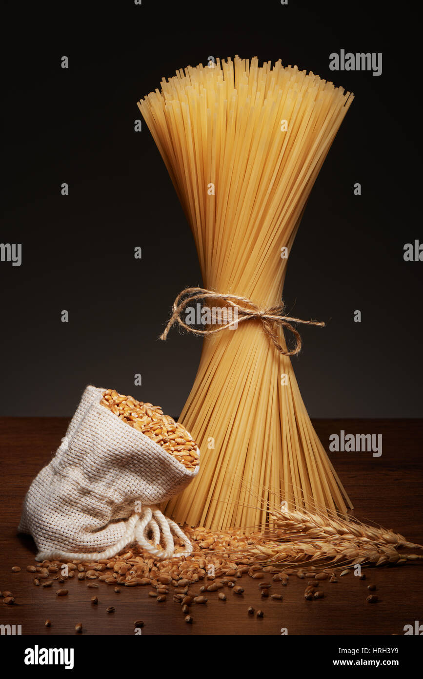 Dry spaghetti pasta tied up with rope, burlap bag of wheat grains and wheat ears on dark wooden table on grey background Stock Photo