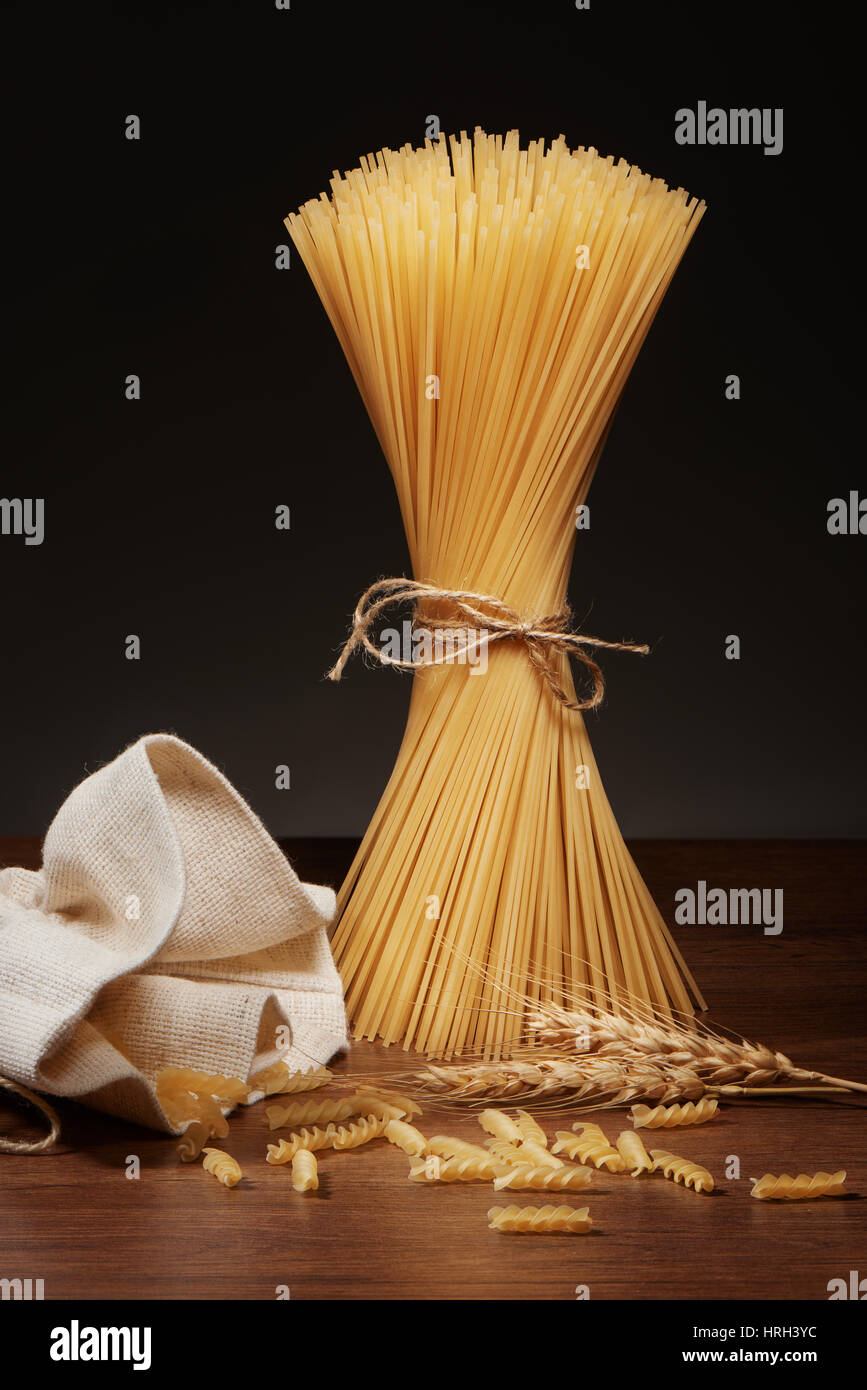 Dry spaghetti pasta tied up with rope, fusilli pasta falling out of burlap bag and wheat ears on dark wooden table on grey background Stock Photo