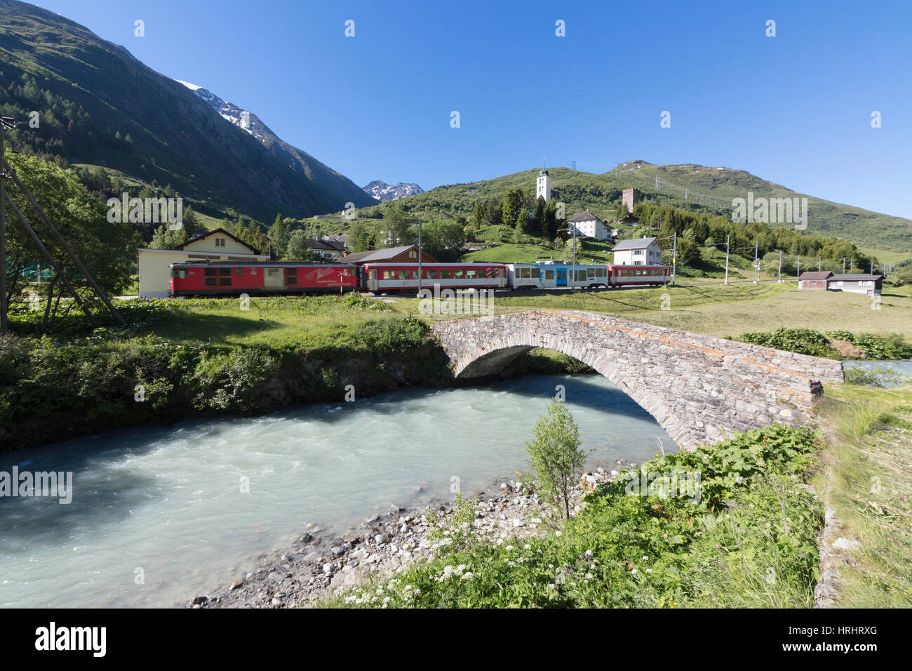 Typical red Swiss train on Hospental Viadukt surrounded by creek and green meadows, Andermatt, Canton of Uri, Switzerland Stock Photo