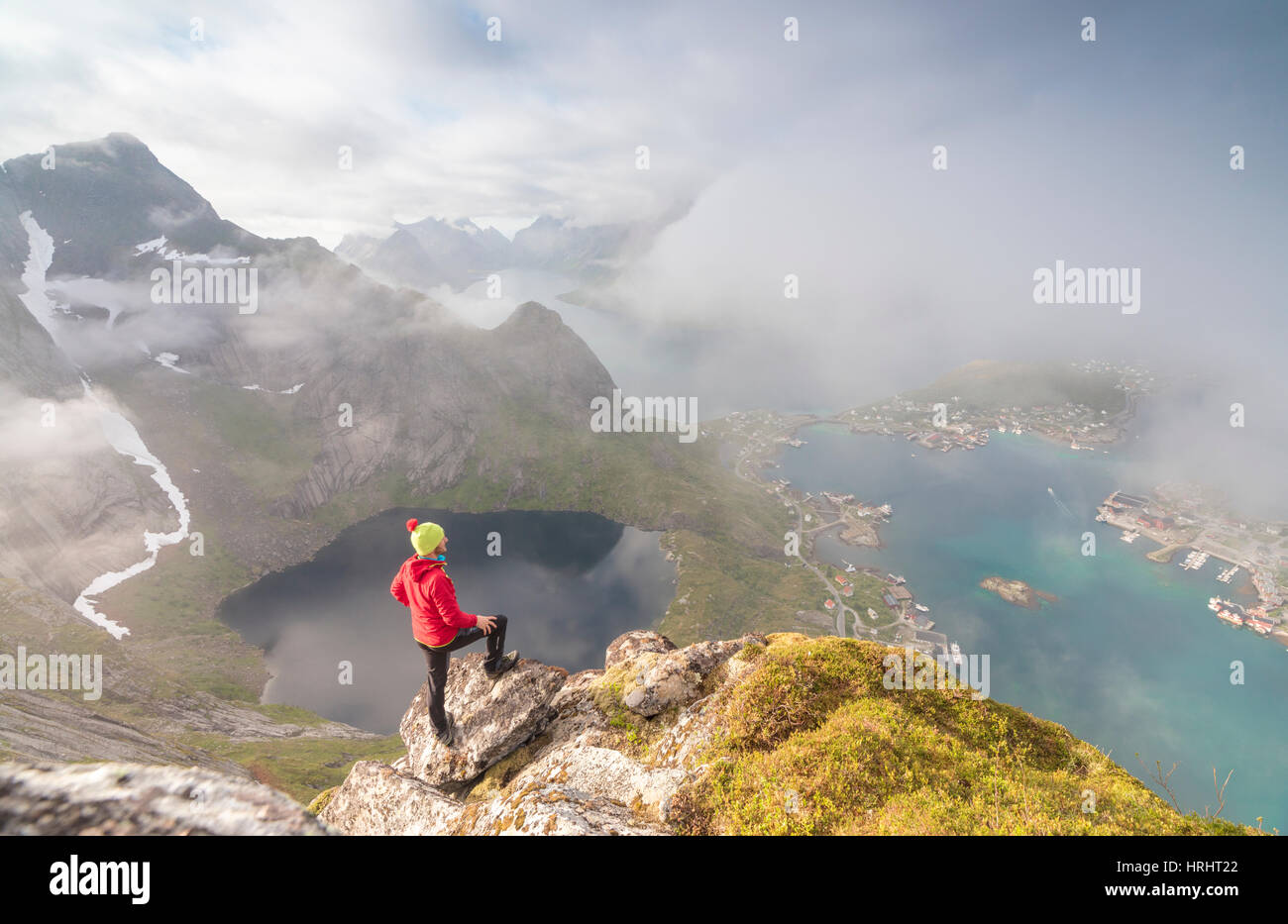 Hiker on summit admires the blue lake and sea framing the village, Reinebringen, Moskenesoya, Lofoten Islands, Norway Stock Photo