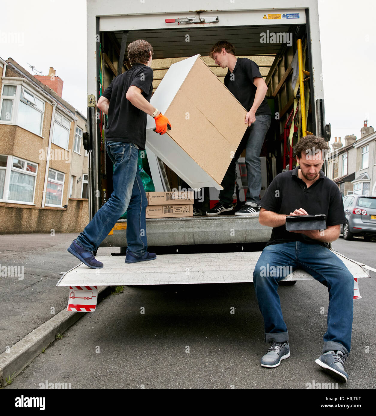 Removals business. A removals company, two men lifting furniture and one seated using a digital tablet. Stock Photo