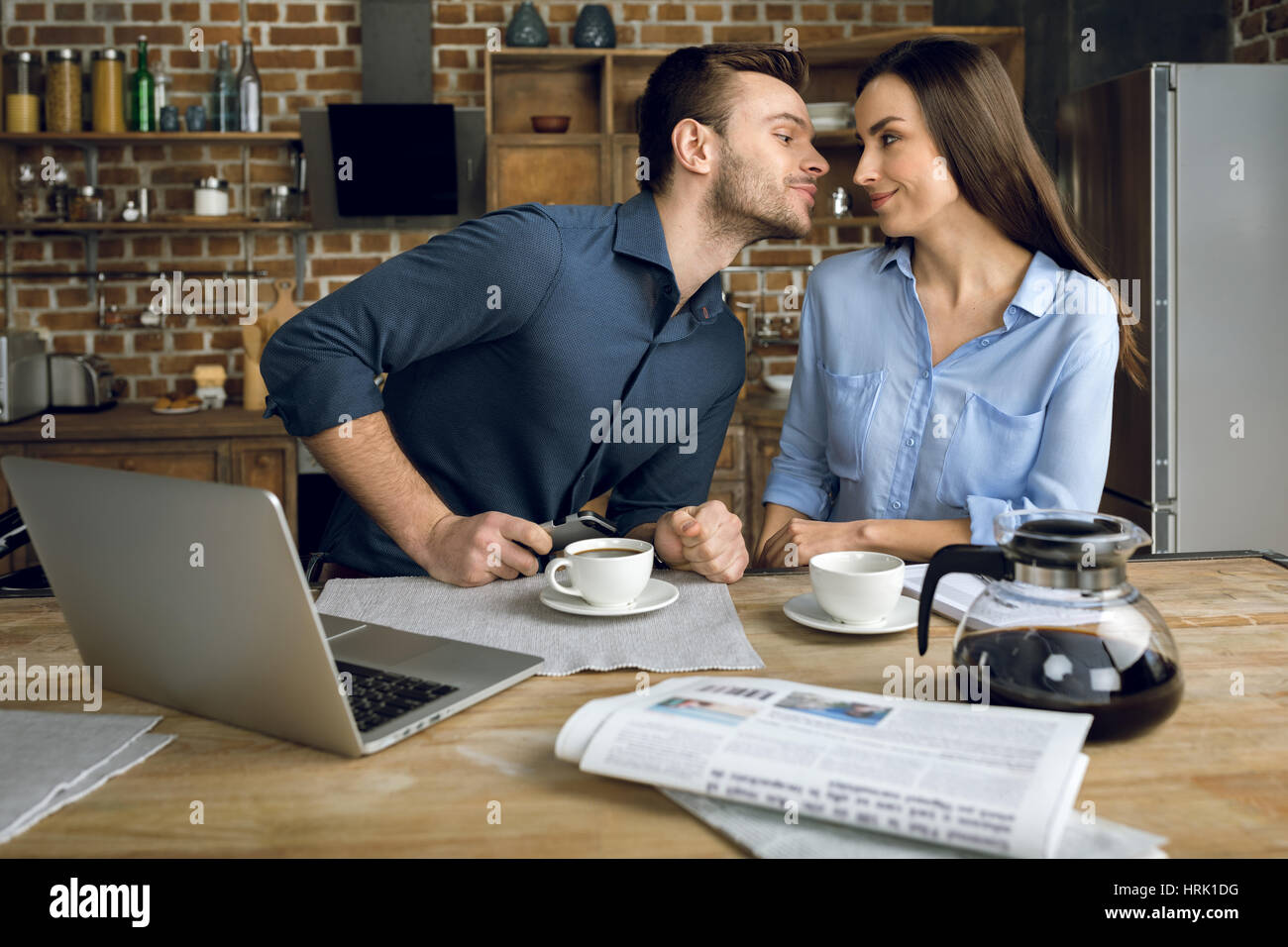 Smiling young couple kissing while drinking coffee in kitchen Stock Photo