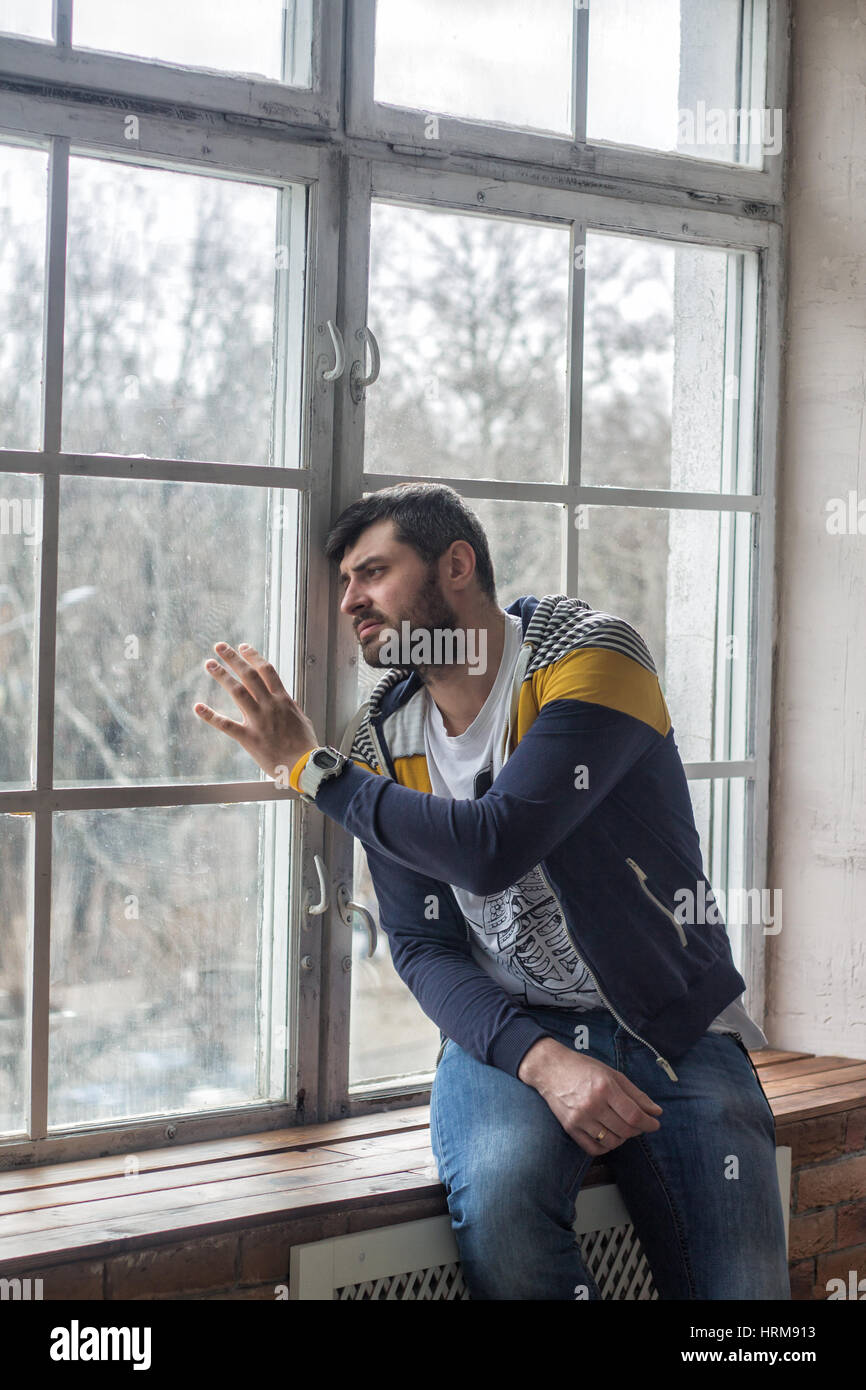 Emotional sad bearded man looking through the window. He is looking worried, depressed, thoughtful and lonely. waiting concept. Stock Photo